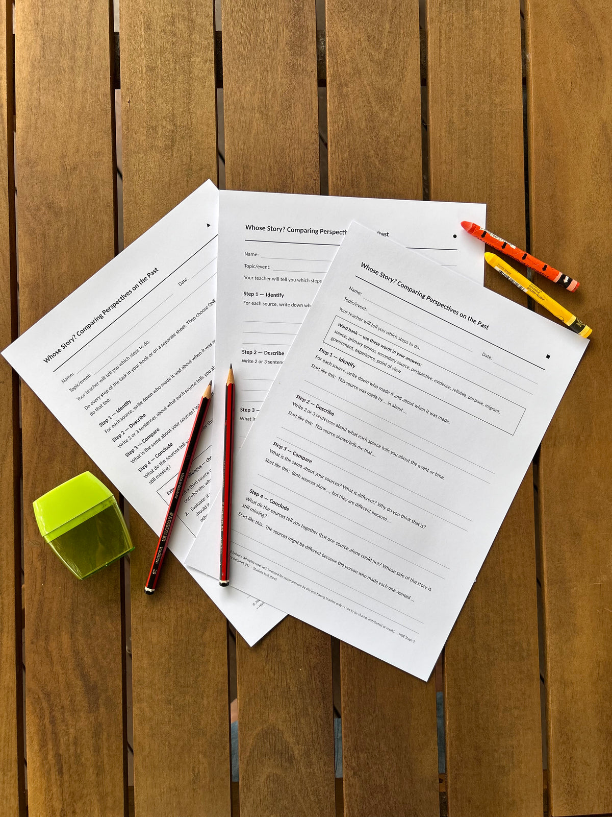 Three versions of the Whose Story? student task sheet spread on a timber table, with pencils, crayons and a sharpener